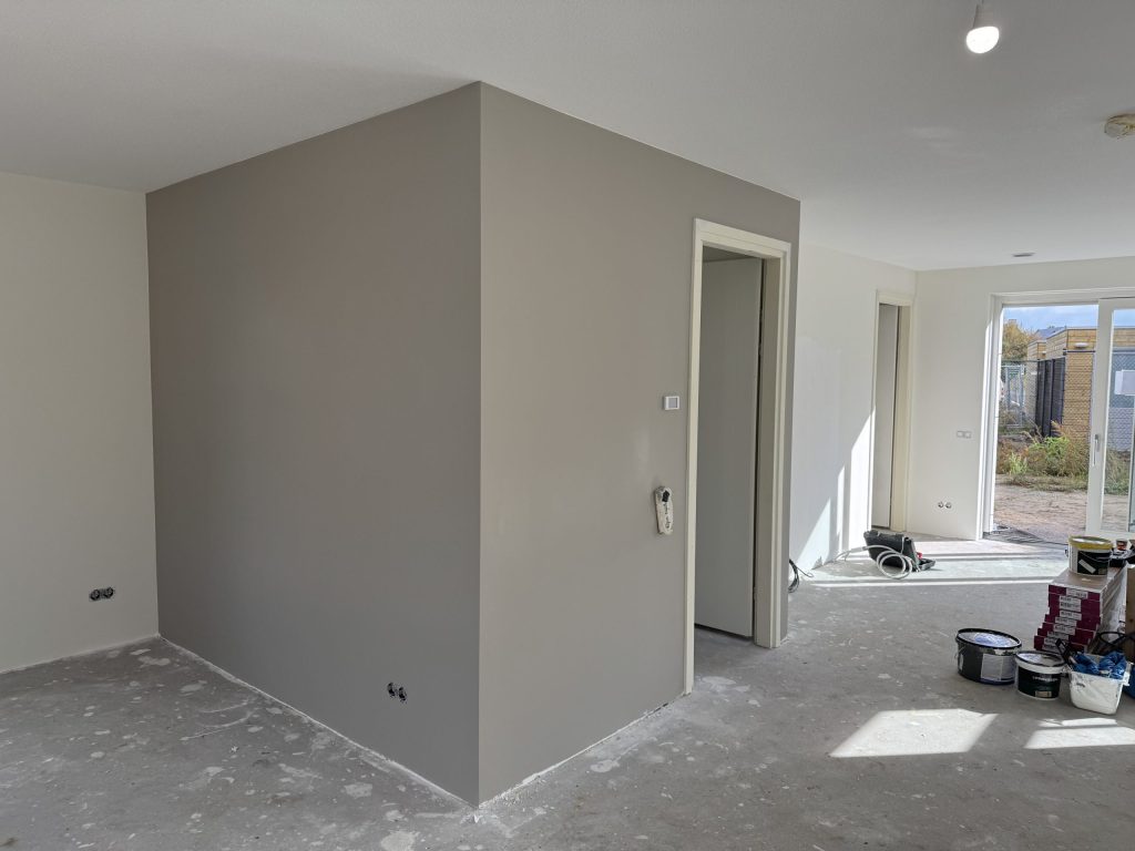 Pas afgewerkte grijze wand rond een deuropening, met verfemmers op de vloer