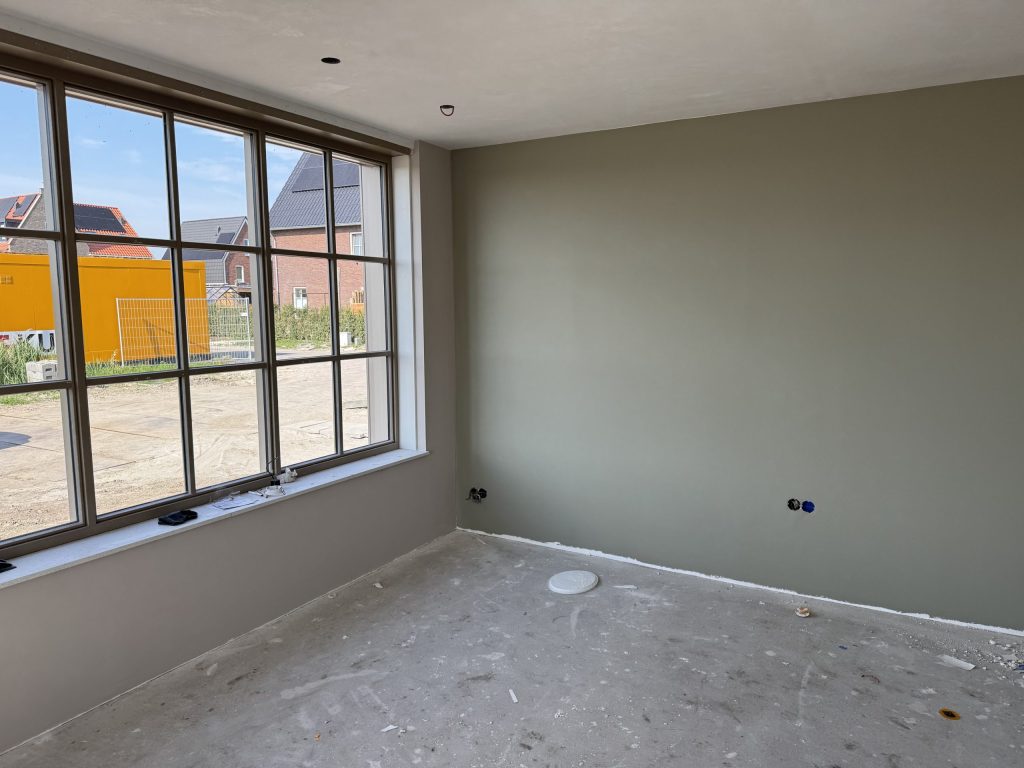 Kamer in een nieuw huis met een groen gesausde wand en een groot raam met uitzicht op de nieuwbouwwijk